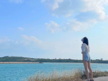 澄みきった水色の海の向こうには島が見えており、海の上にはきれいな青色の青空が広がっている。その海辺で、白いシャツを着てデニムの短パンを履いた一人の女が、空を見上げている。女の髪は風になびいている。