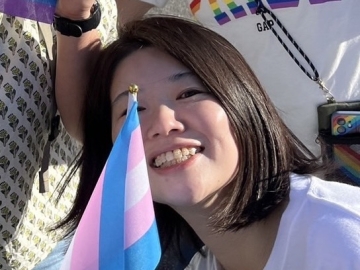 岩﨑さんのプロフィール写真：笑顔の岩﨑さんが小さなトランスプライドフラッグを顔の前に掲げている。