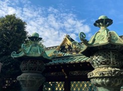空を背景に、手前に銅製の古風な灯篭と奥に日本の寺社(上野東照宮)の屋根が見える写真。手前の灯篭には繊細な彫刻と徳川将軍家の葵の御紋が施されており、背後の建物には青緑色の屋根瓦と金色の装飾が施されています。左側には木々も映っています。