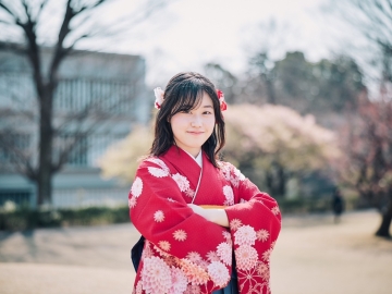 ミディアムの⻑さの髪の⽑で、⾚い着物と紺の袴を着たさりが、笑顔で腕を組んで⽴っている。背景は、卒業式の⽇の春のキャンパスの⽊々。