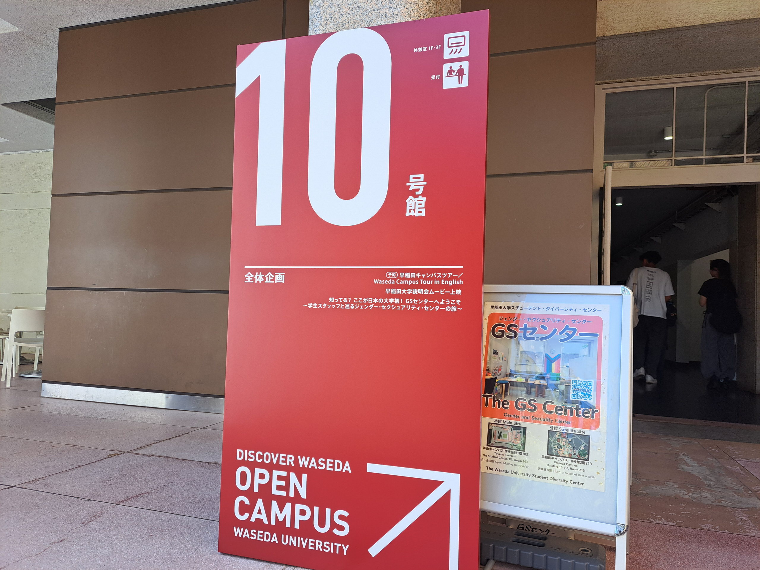 早稲田大学10号館1階入り口に設置された大型の立て看板と案内ポスターの様子を写した写真。左側に大きく目立つ赤い縦長の看板があり、上部に白抜きで「10号館」と横書きで大きく表示されている。その下に「全体企画」として、いくつかのプログラム名が小さめに横書きで掲載されている。プログラム名としては上から順に「早稲田キャンパスツアー/Wseda Campus Tour In English 」、「早稲田大学説明会ムービー上映」、「知ってる?ここが日本の大学初!GSセンターへようこそ~学生スタッフと巡るジェンダー·セクシュアリティ·センターの旅~」などの三つのものが横書きで書かれている。看板の下部には白い英語で「DISCOVER WASEDA OPEN CAMPUS WASEDA UNIVERSITY」と書かれ、 右側には、さらに小さめのポスター掲示板があり、「GSセンター」の文字が大きく見える。ポスターにはカラフルなイラストや写真が散りばめられており、上部に「スチューデント・ダイバーシティ・センター」の表記が横書きで、黒字で書かれている。さらにポスター中央よりやや下側に歯「The GS Center」と大きく英語で表記され、その下に「Gender and Sexuality Center」の英語表記もある。ポスター中央部右側にはQRコードがある。 背景には建物の出入口があり、2人の来場者が奥に歩いていく様子が見える。床は淡いピンクがかったタイル張りで、壁面は茶色とベージュの色調。