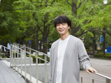 谷口洋幸さんの写真。白いTシャツにグレーのカーディガンを羽織った谷口さんが、スロープの手すりに左手を添えながら立っている写真。背景に、葉が生い茂った並木が写っている。